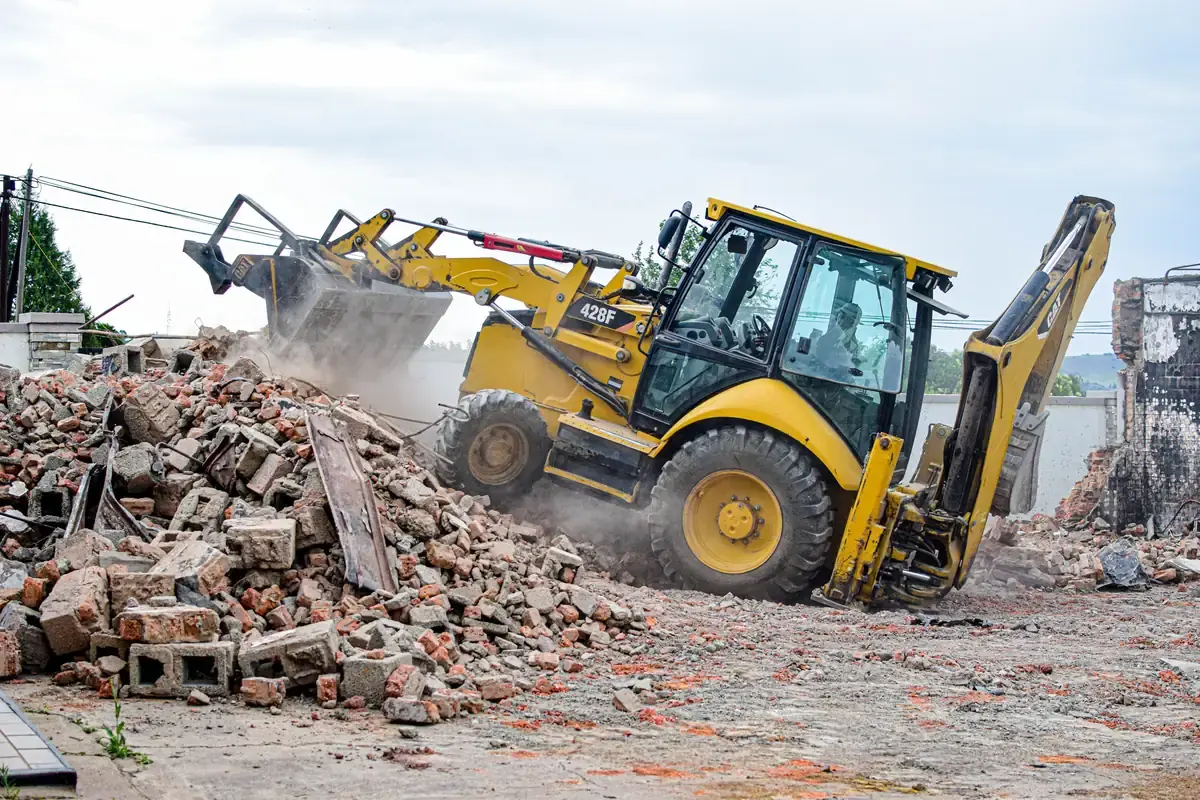 A backhoe loader pushing through a pile of brick rubble, dust rising around the wheels