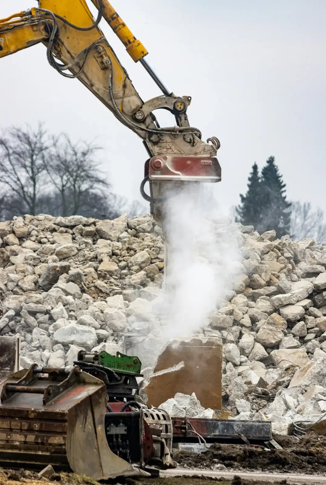 An excavator breaker throwing up a plume of dust as it works into concrete rubble