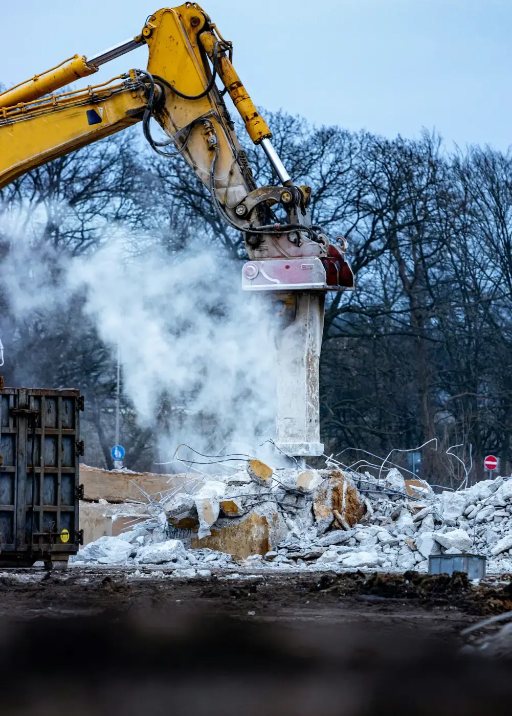 An excavator breaker at work on concrete, with a waste skip standing alongside