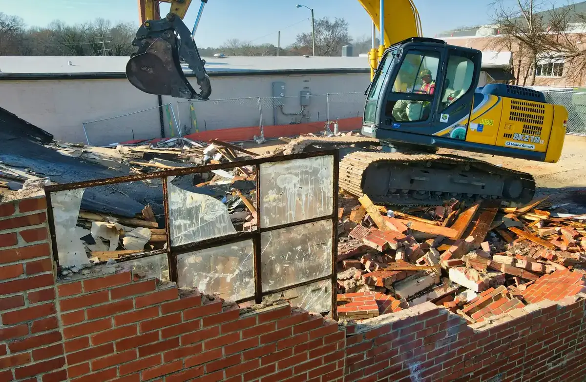 An excavator working on the roof of a brick building, a steel-framed window in the foreground