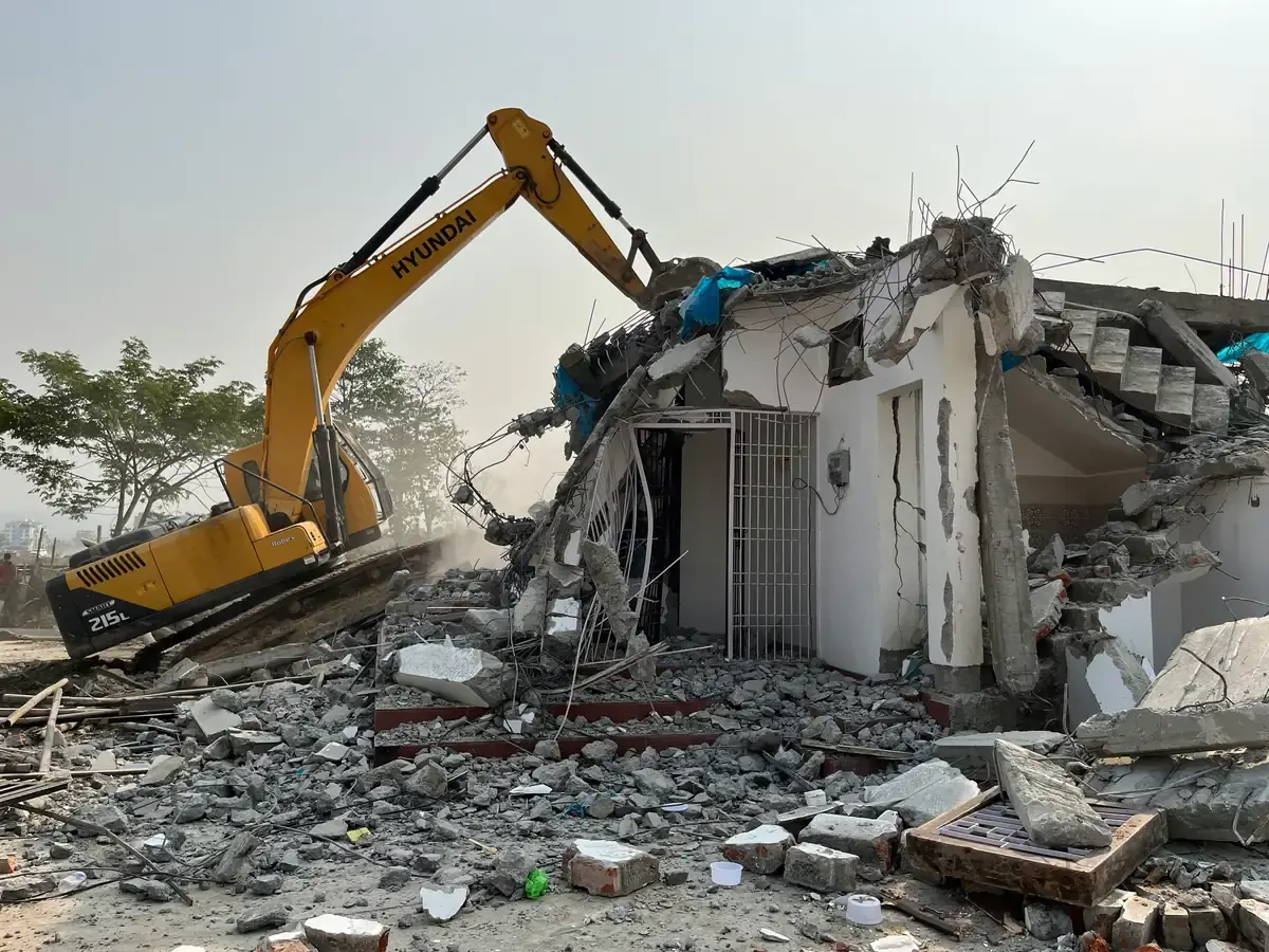 An excavator breaking a concrete house, exposed reinforcement bars twisting out of the wall