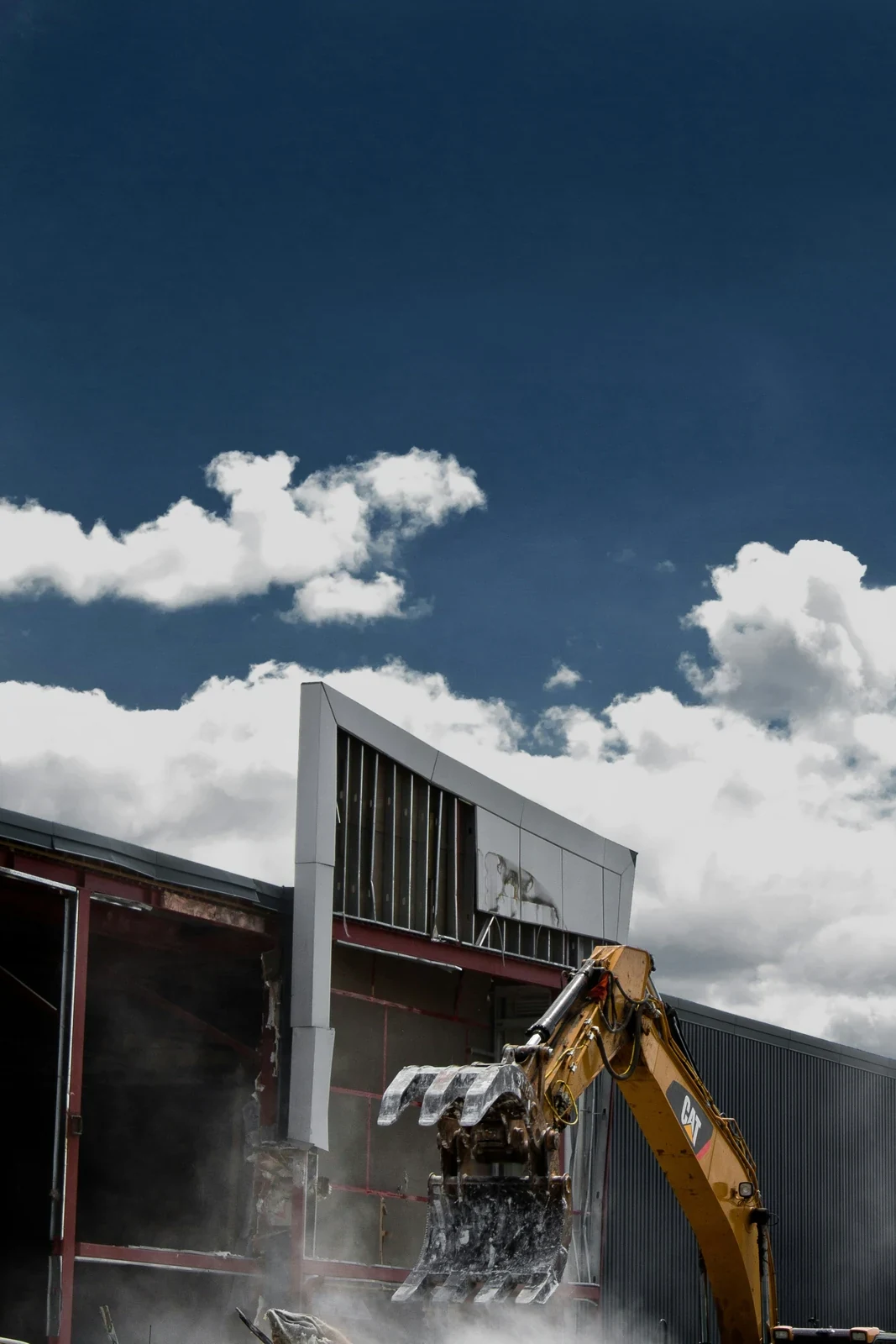 An excavator with a grapple pulling cladding from a metal-clad industrial building