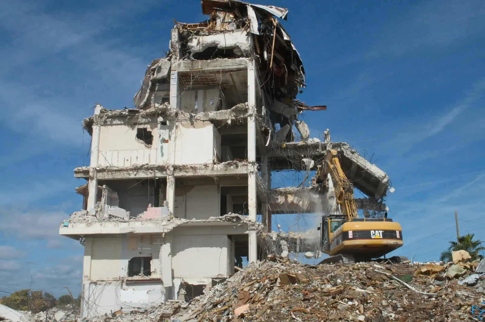 An excavator standing on a rubble platform to reach a multi-storey concrete building