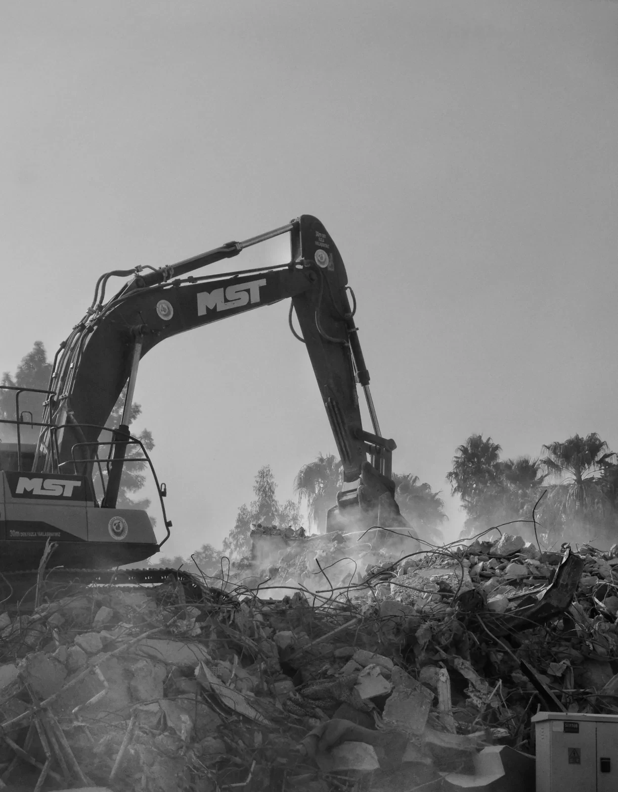 A monochrome view of an excavator working a rubble field with dust hanging in the air