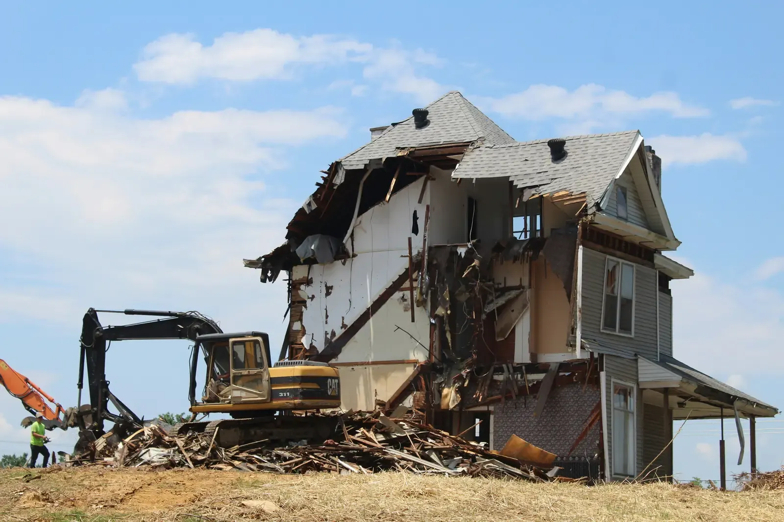 An excavator taking down a two-storey timber house, framing and boards in the bucket