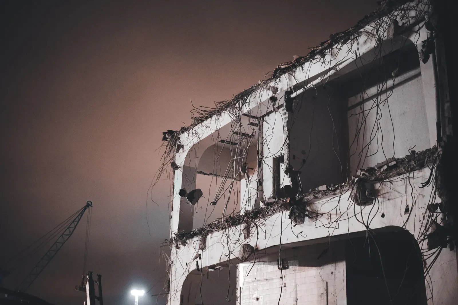 A part-demolished floor slab at night, cables hanging from the cut edge and a crane behind