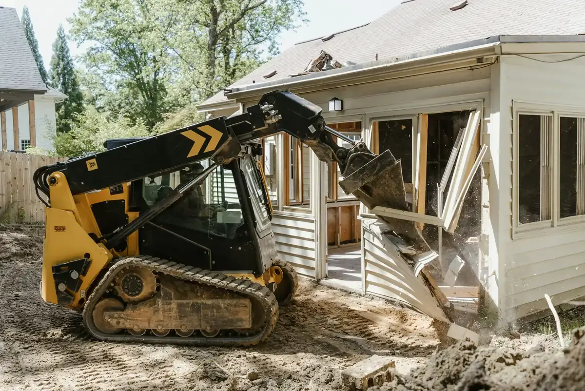 A compact track loader pulling siding from the sunroom of a house