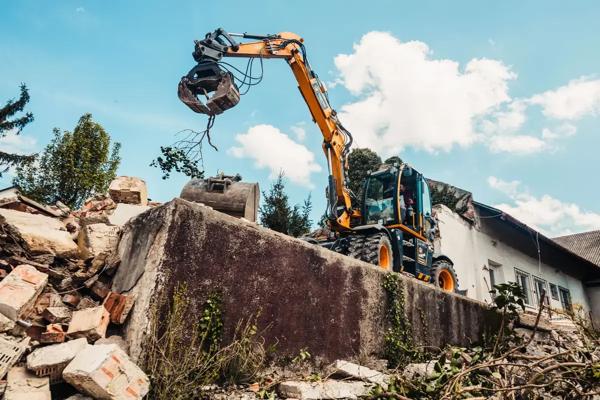 A wheeled excavator reaching over a concrete wall with a grapple, rubble in the foreground